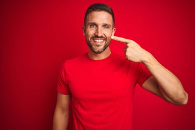 Kırmızı izole edilmiş arka plan üzerine gündelik tişört giyen genç yakışıklı bir adam. El parmağı ve burunla işaret ediyor. Neşeli gülümsüyor. Güzellik konsepti