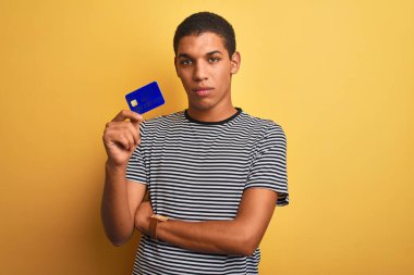 Genç, yakışıklı, Arap bir adam elinde kredi kartıyla izole edilmiş sarı arka planda duruyor. Akıllı bir yüz ifadesiyle ciddi olduğunu düşünüyor.
