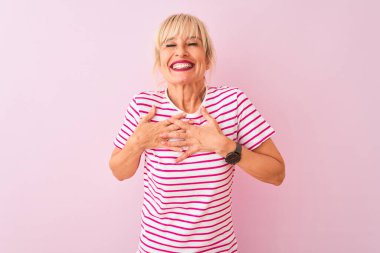 İzole edilmiş pembe arka planda duran çizgili tişört giyen orta yaşlı bir kadın. Elleri göğsünde, gözleri kapalı ve yüzünde minnet dolu bir jest ile gülümsüyor. Sağlık kavramı.