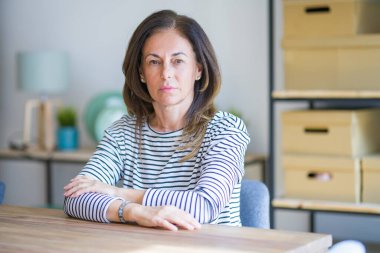 Orta yaş yaşlı kadın yüzünde ciddi ifade ile evde masada oturan. Kameraya basit ve doğal bir bakış.