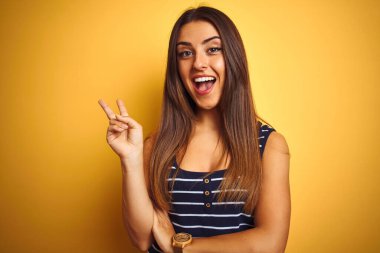 İzole sarı arka plan üzerinde duran çizgili t-shirt giyen genç güzel kadın mutlu yüz ile gülümseyerek zafer işareti yapıyor kameraya göz kırparak. İki numara.