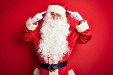 Noel Baba kostümü giymiş orta yaşlı yakışıklı bir adam izole edilmiş kırmızı arka planda dikiliyor iki eliyle başını gösteren bir gülümseme, harika bir fikir ya da düşünce, iyi bir hafıza.