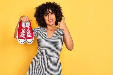 Kıvırcık saçlı genç bir Arap kadın izole edilmiş sarı arka planda spor ayakkabıları tutuyor. Gülümsüyor ve gülümsüyor. İşaret veriyor.
