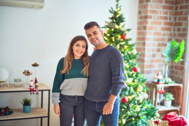 Mutlu ve kendinden emin gülümseyen genç bir çift. Evde Noel ağacının etrafında dikilip sarılıyorsun.