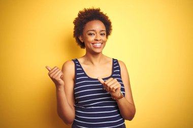 Beauitul Afro-Amerikalı kadın izole edilmiş sarı arka planda yaz tişörtü giyiyor. Arka tarafı işaret ediyor. El ve başparmakları havada, kendinden emin.