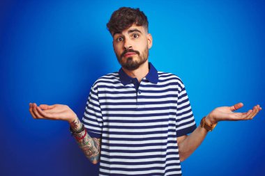 İzole edilmiş mavi arka planda duran çizgili polo giyen genç adam elleri ve kolları havada, kafası karışmış bir ifade takınıyor. Şüphe kavramı.