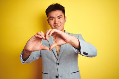 Genç Asyalı Çinli işadamı, izole edilmiş sarı arka planda duran ceketini giymiş ve elleriyle kalp sembolünü ve şeklini gösteren aşkla gülümsüyor. Romantik konsept.