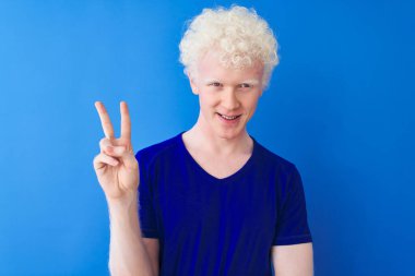 Sıradan tişörtler giyen genç, sarışın bir adam izole edilmiş mavi arka planda dikiliyor ve kameraya gülümsüyor zafer işareti yapıyor. İki numara..