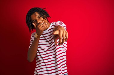 Rastalı tişörtlü genç afro adam izole edilmiş kırmızı arka planda dikiliyor sana gülüyor, eli ağzında kamerayı işaret ediyor, utanç ifadesi takınıyor.