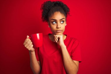 Genç Afro-Amerikan kadın, izole edilmiş kırmızı arka planda bir fincan kahve içiyor. Ciddi bir yüz. Soru üzerine kafa karıştırıcı bir fikir.