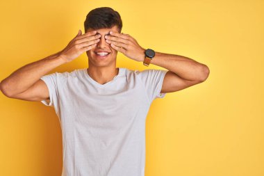 İzole edilmiş sarı arka plan üzerinde duran beyaz t-shirt giyen genç Hintli adam neşeli ve komik gülümseyen elleriyle gözlerini kapatıyor. Kör kavram.