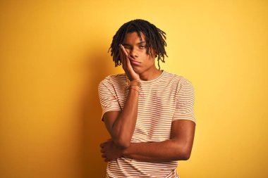 Sarı arka plan üzerinde duran çizgili t-shirt giyen Afro bir adam kollarını kavuşturarak depresyondan sıkılmış ve yorgun göründüğünü düşünüyor..