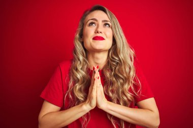 Basit bir tişört giyen genç ve güzel bir kadın kırmızı izole edilmiş arka planda duruyor. Elleriyle yalvarıyor ve dua ediyor. Yüzünde umut ifadesi var. Af diliyor. Din kavramı.