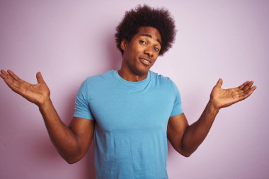 Afro saçlı, mavi tişörtlü, soyutlanmış pembe arka planda duran, elleri ve kolları havada, kafası karışmış Afro-Amerikan bir adam. Şüphe kavramı.