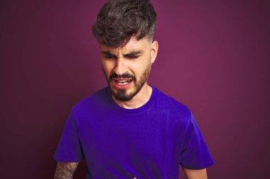 Dövmeli genç bir adam, midesi ağrıyan, hazımsızlık çeken ve kendini kötü hisseden mor arka plan üzerinde duruyor. Ache kavramı.