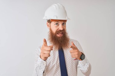 İzole edilmiş beyaz arka plan üzerinde güvenlik kaskı takan genç kızıl saçlı mimar mutlu ve komik yüzlü parmaklarını kameraya doğrultuyor. İyi enerji ve titreşimler.