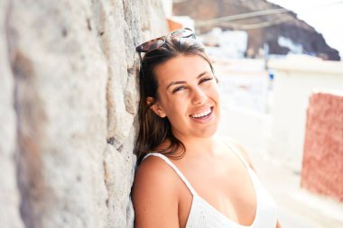 Güzel kız beyaz duvara yaslanıyor, genç arkadaş canlısı kadın mutlu bir yaz gününde gülümsüyor.