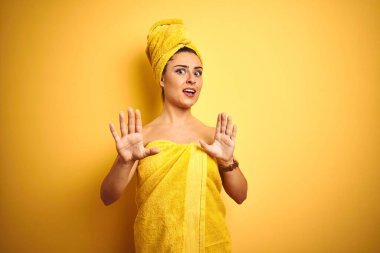 Young beautiful woman wearing towel after shower over isolated yellow background afraid and terrified with fear expression stop gesture with hands, shouting in shock. Panic concept.