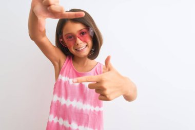 Pembe tişört ve güneş gözlüğü takan güzel bir kız çocuğu izole edilmiş beyaz arka planda gülümsüyor ve mutlu yüzlü elleri ve parmakları olan bir çerçeve yapıyor. Yaratıcılık ve fotoğrafçılık kavramı.