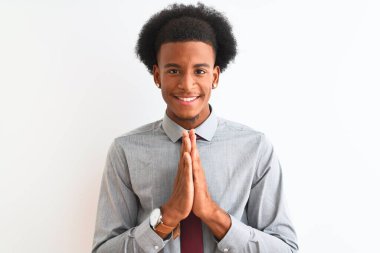 Genç Afro-Amerikalı iş adamı kravatını takmış, beyaz arka planda durmuş ellerini birleştirip af diliyor, kendinden emin bir şekilde gülümsüyor..