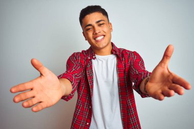 Kırmızı tişörtlü genç adam, izole edilmiş beyaz arka planda dikilip kameraya bakarak sarılmak için kollarını açmış gülümsüyor. Mutluluğu kucaklayan neşeli bir ifade..