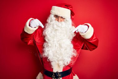 Orta yaşlı, yakışıklı, Noel Baba kostümü giymiş, izole edilmiş kırmızı arka plan üzerinde duran üzgün ve üzgün görünen, parmaklarıyla yönü gösteren, mutsuz ve depresif..