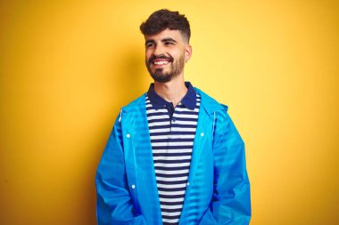 Yağmurluk giyen genç yakışıklı adam izole edilmiş sarı arka plan üzerinde duruyor yüzünde gülümseme, doğal bir ifade ile yan yana bakıyor. Kendinden emin gülen.