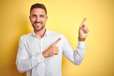 Sarı izole edilmiş arka plan üzerinde zarif beyaz gömlek giyen genç yakışıklı adam gülümsüyor ve kameraya bakıyor iki eliyle ve parmaklarıyla kenarı işaret ediyor..