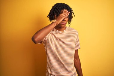 Rasta desenli tişörtlü Afro adam izole edilmiş sarı arka planda duruyor gözlerini eliyle kapatıyor, ciddi ve üzgün görünüyor. Görme, saklama ve reddetme konsepti