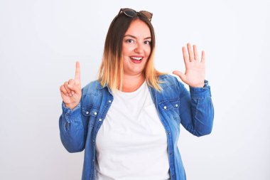 İzole edilmiş beyaz arka plan üzerinde duran kot gömlek giyen güzel bir kadın altı numaralı parmaklarını gösterip işaret ederken kendinden emin ve mutlu gülümsüyor..