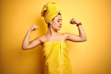 İzole edilmiş sarı arka planda duş üstüne havlu giyen güzel bir kadın gururla gülümseyen kol kaslarını gösteriyor. Fitness konsepti.