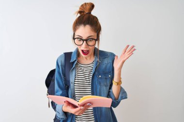 Sırt çantası giyen kızıl saçlı bir kadın, izole edilmiş beyaz arka plan üzerine kitap okuyor. Çok mutlu ve heyecanlı, kazanan yüz ifadesi zaferi büyük bir gülümsemeyle kutluyor ve ellerini kaldırıyor.