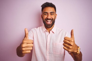 İzole edilmiş pembe arka plan başarısı tabelasının üzerinde dikilen rahat gömlekli genç Hintli adam elleriyle olumlu jest yapıyor, gülümsüyor ve mutlu. Neşeli ifade ve kazanan jest.