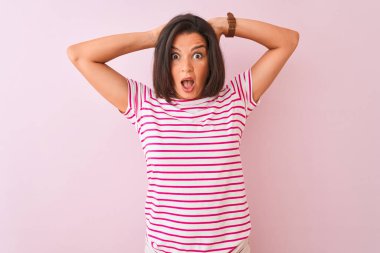 İzole edilmiş pembe arka plan üzerinde duran çizgili tişört giyen genç ve güzel bir kadın elleri kafasında çılgın, korkmuş ve ağzı açık şoktan şaşırmış.