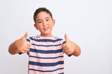 Güzel çocuk, izole edilmiş beyaz arka planda dikilmiş sıradan çizgili tişört giyiyor. Elle olumlu jest yapıyor, gülümsüyor ve başarı için seviniyor. Kazanan jest.
