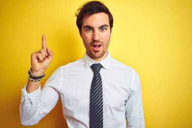 Zarif gömlek ve kravat giyen genç yakışıklı iş adamı izole edilmiş sarı arka plan üzerinde başarılı bir fikirle işaret ediyor. Heyecanlı ve mutlu. Bir numara..