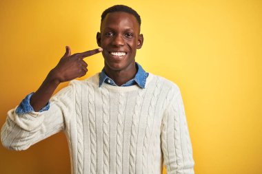 İzole edilmiş sarı arka plan üzerinde kot gömlek ve beyaz kazak giyen Afro-Amerikan bir adam. El parmağı ve burunla işaret ediyor. Neşeli gülümsüyor. Güzellik konsepti