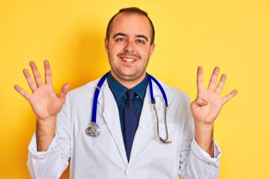 Paltolu ve steteskoplu genç doktor izole edilmiş sarı arka planda duruyor ve dokuz numaralı parmaklarıyla işaret ederken kendinden emin ve mutlu bir şekilde gülümsüyor..