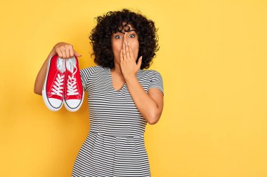 Kıvırcık saçlı genç bir Arap kadın izole edilmiş sarı arka planda spor ayakkabıları tutuyor. Ağzı kapalı. Yanlışlıkla utançla dolu.