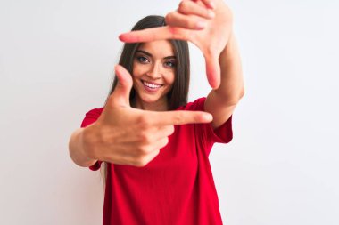 İzole edilmiş beyaz arka planda kırmızı bir tişört giyen genç ve güzel bir kadın gülümsüyor ve elleri ve parmakları mutlu yüzlü bir çerçeve yapıyor. Yaratıcılık ve fotoğrafçılık kavramı.