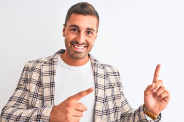 Zarif bir ceket giyen genç yakışıklı iş adamı izole edilmiş arka planda gülümsüyor ve kameraya bakıyor iki eliyle ve parmaklarıyla kenarı işaret ediyor..