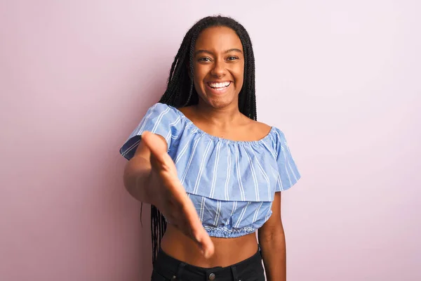 African american woman wearing striped t-shirt standing over isolated pink background smiling friendly offering handshake as greeting and welcoming. Successful business.