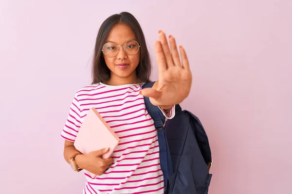 Chinese student woman wearing glasses backpack holding book over isolated pink background with open hand doing stop sign with serious and confident expression, defense gesture