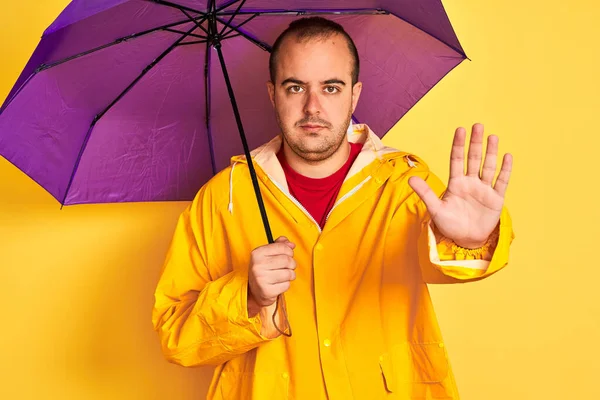 Young man wearing raincoat holding purple umbrella standing over isolated yellow background with open hand doing stop sign with serious and confident expression, defense gesture