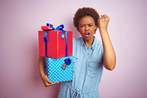 African american woman holding birthday gifts over pink isolated background annoyed and frustrated shouting with anger, crazy and yelling with raised hand, anger concept