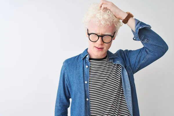 Young albino blond man wearing denim shirt and glasses over isolated white background confuse and wondering about question. Uncertain with doubt, thinking with hand on head. Pensive concept.