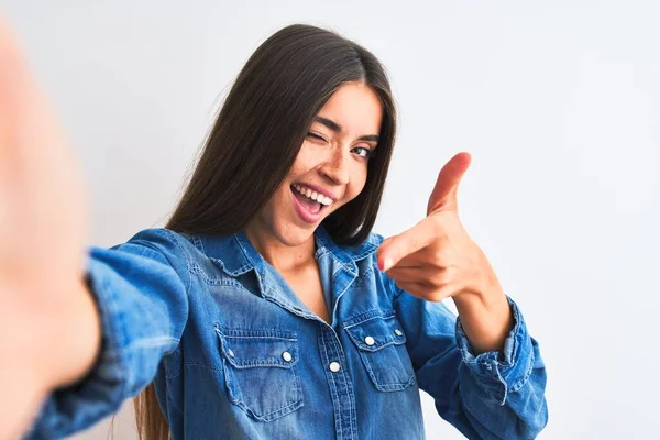 Beautiful woman wearing denim shirt make selfie by camera over isolated white background pointing fingers to camera with happy and funny face. Good energy and vibes.