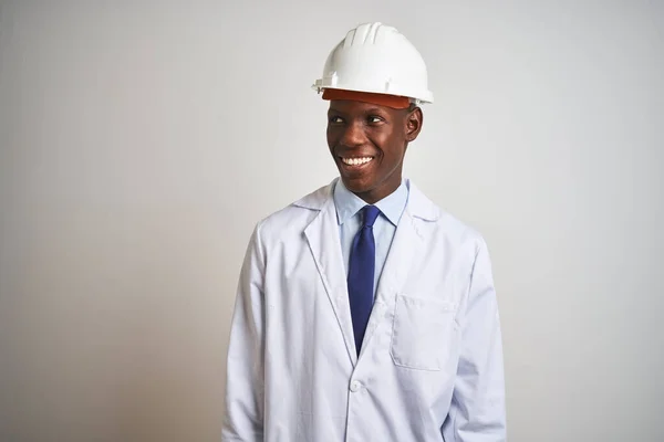 African american engineer man wearing coat and helmet over isolated white background looking away to side with smile on face, natural expression. Laughing confident.