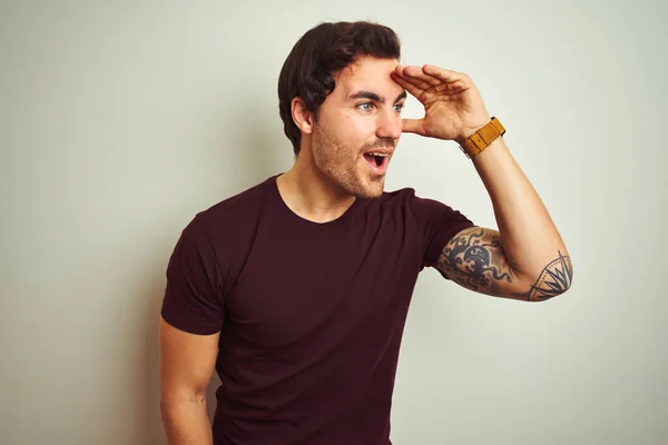 Young handsome man with tattoo wearing purple casual t-shirt over isolated white background very happy and smiling looking far away with hand over head. Searching concept.