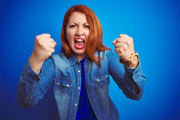 青い隔離された背景の上にデニムシャツを着た若い美しい赤毛の女性は怒りで叫んでいる間に怒りと怒りに苛まれ激怒拳を上げ怒っています 激怒と積極的な概念 — ストック写真
